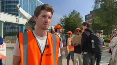 A man wearing an orange reflective vest with a white T-shirt underneath stands outside a hospital. Others can be seen behind him holding placards and wearing orange hats.