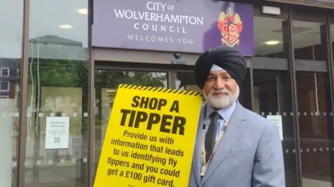 City of Wolverhampton Council Councillor Bhupinder Gakhal, cabinet member for resident services, has a short white beard and wears a light grey suit, light blue shirt, black tie and a black turban. He is holding a big yellow sign that reads, 'Shop a tipper, provide us with information that leads to us identifying fly tippers and you could get a £100 gift card'.
