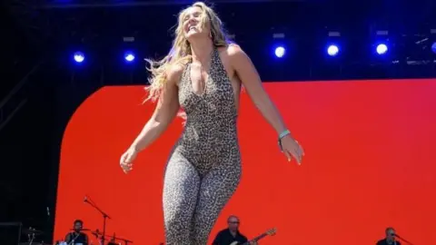 Lauren Filer in a leopard print costume on a big stage with a red background.   There are men playing instruments behind her.
