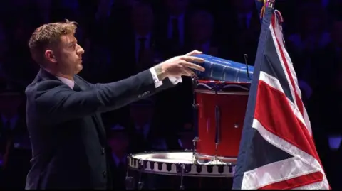 A man wearing a suit places a blue book on top of a drum, next to the Union Flag