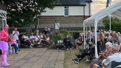 A man stood holding a microphone while stood on a patio. He is speaking to a large group of people sat beneath a white gazebo