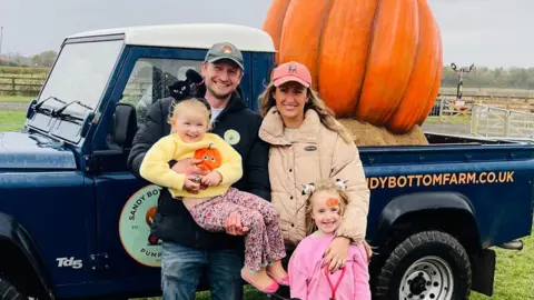 A man, Chris Sadler, stands next to his wife and two children. They are standing on a farm, in front of a blue truck with a large pumpkin sculpture in the back. Chris Sadler is wearing blue jeans, a black hooded coat and a grey cap. He is holding a young girl who is wearing pink shoes, colourful trousers and a yellow sweater with a pumpkin on. His wife is wearing a beige coat with a pink cap on. Another young girl is standing in front of her wearing a pink sweater. 