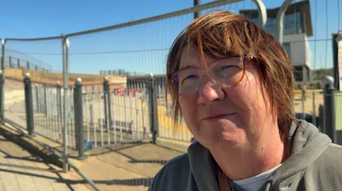 A woman wearing brown horn rimmed glasses looks at the camera while standing in front of metal fencing surrounding the Sutton paddling pool