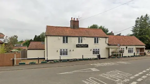 Google Roadside view of Fox and Hounds pub, a cream double-storey building with a single-storey extension. It is located on the side of a road. 
