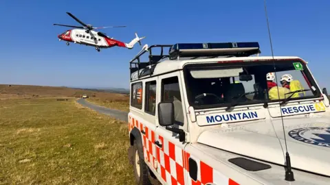 North Dartmoor Search and Rescue Team The coastguard helicopter is taking off on Dartmoor on a bright blue sky day. A Mountain Rescue-branded Land Rover is parked in the foreground. at the end of a winding lane another emergency vehicle is parked and surrounded by uniformed people.