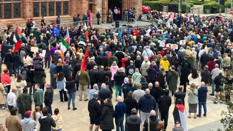 crowds with colourful flags, facing the Guildhall with speakers set up at the front 