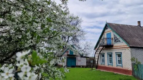 Vladyslava Bondar A detached brick house in the countryside with turquoise blue window frames. A tree in bloom is in the foreground, another out building can be seen in the background. 