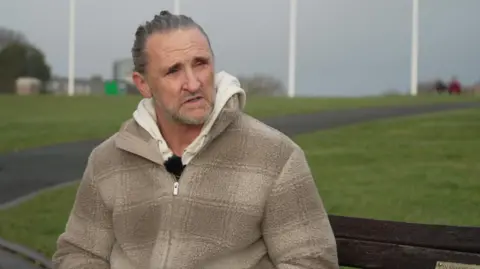 Paul Maxwell, wearing a beige fleece and hoodie, looks towards the right of frame. He is sat on a bench on Plymouth Hoe.