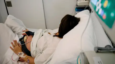A generic photo of a woman lying in a hospital bed facing away from the camera. She is hooked up to several drips and a monitoring screen, with a black band around her stomach. She is wearing a white gown and resting on white pillows.