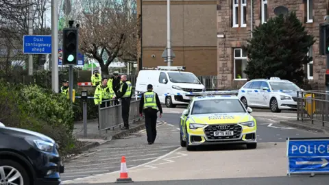 Stuart Cowper Police officers walk on a pavement. The road has been blocked by a police car and police tape and a cone. A white car and van can be seen behind the police car