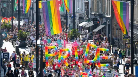 Pride Cymru supporters parade through the city centre in Cardiff. Rainbow balloons spell PRIDE and attendees line the streets. 