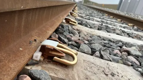 BBC Close-up image of metal railway tracks, metal bolt fasteners, concrete supports and large pieces of gravel.