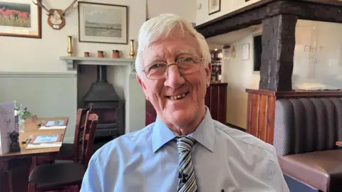 Eric Norman, a man in his 70s wearing glasses, a blue shirt and striped tie, is sat in his pub with tables, a fire and pictures on the wall behind him