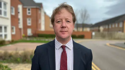 Profile picture of Paul Bristow. He wears a dark jacket, a white pinstripe shirt and burgundy spotty tie. In the background are some blurred-out houses.