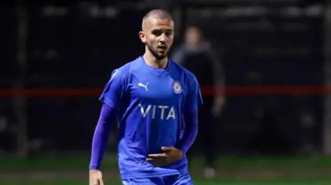 Bader Al-Akkad Bader Al-Akkad wears a blue Stockport County kit while standing with a football on the pitch during a match.
