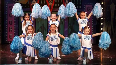 Supplied Six girls line up on a stage. They are all dressed in white and blue cheerleader outfits and are holding blue and white pompoms. All six have their hair in bunches with blue and white ribbons. 