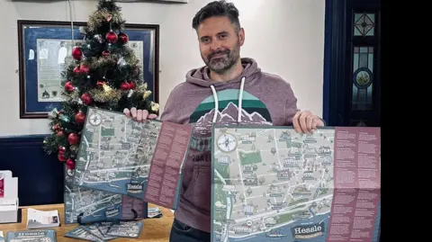 Richard Royal pictured wearing a lilac coloured hoodie, with a decorated Christmas tree to his left, holding two difference sizes of his local history tour map.