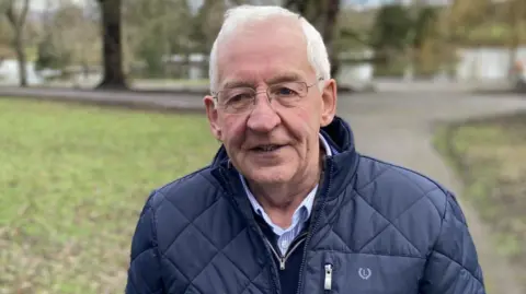 Les McCaffrey wears a navy quilted coat with a navy jumper and a navy shirt on underneath. He has short silver hair and wears clear-framed glasses. he is standing in a park which has green trees, grass and a pond.