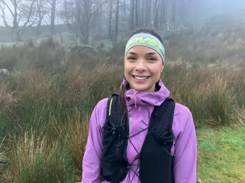 Sarah Perry. She is smiling and wearing a black running bag over a purple jacket. Her brown hair is tied back and she is wearing a headband. Behind her are trees and grassy bushes.