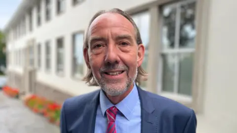 Deputy Steve Falla, a man with shoulder length brown hair and a short grey beard. He is wearing a blue suit and a blue shirt with a red striped tie. 
