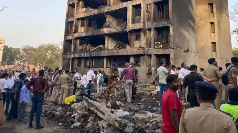 A large crowd of people stand around piles of rubble at the crash site. A tall building, although still standing, has been destroyed by the crash. It is charred black and has suffered extensive damage.