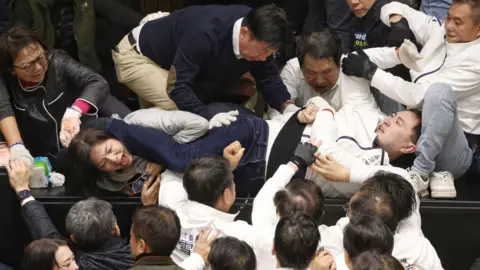 Legislators from various political parties wearing dark or light coloured jackets brawl in the Taiwan legislature, with several people trying to pull apart other people in the middle physically fighting with each other