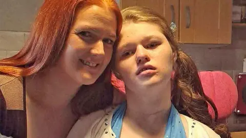Claire McGill is pictured beside her daughter Sianon who is in a wheelchair. Their heads are touching as they pose for the photo. Claire has red hair and is smiling. Sianon has her hair tied back and is wearing a white top with a light blue scarf.