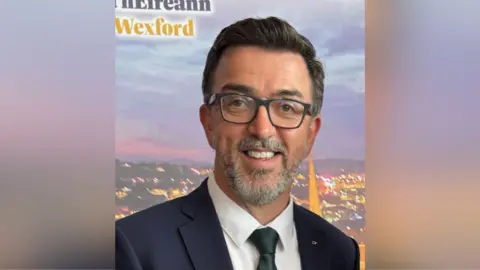 Brendan McAleer Brendan McAleer. He has hark hair and a grey beard, wearing a navy blazer, white shirt and dark green tie.