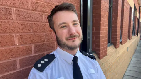 Ch Insp Chris Tinney stands close to a red brick wall outside a building, wearing police uniform. A brick wall runs behind him and the building has a row of windows with paving outside.
