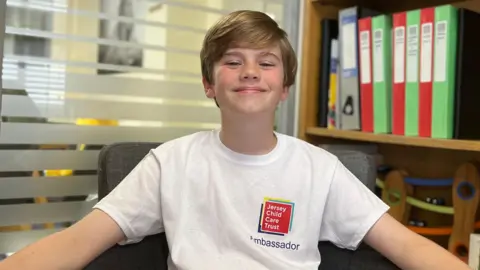 BBC Henry is wearing a white “Jersey Child Care Trust Ambassador” T-shirt sits in a chair with arms resting outward, in an office setting with shelves of folders, books, and small objects in the background.