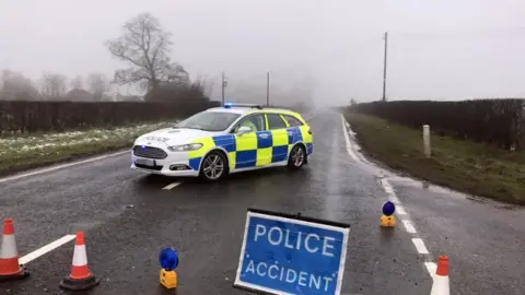 Pacemaker A yellow, blue and white police car parks side ways on a road which is wet with snow on the grass at the side. There is a blue 'police accident sign' and orange and white cones. The sky is grey with fog.