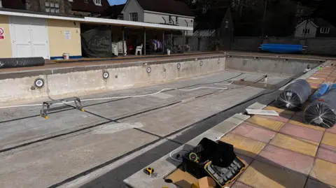 The empty pool before it was refurbished. It is grey and looks tired. There is a set of tools on the side of the pool and a workbench in it.