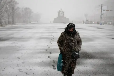 Kim Hong-ji/Reuters Seorang wanita terbungkus cuaca musim dingin dengan mantel tebal dan syal di kepalanya berjalan melalui jalan yang sepi ketika salju turun dan menyelimuti tanah. Ada sebuah monumen di kejauhan di belakangnya.