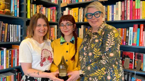 Three women are standing in front of a colourful bookshelf holding a golden trophy. 