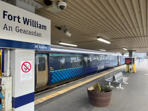 A blue train on a platform, with a sign reading "Fort William" in the foreground. Benches and plants can be seen on the platform. 