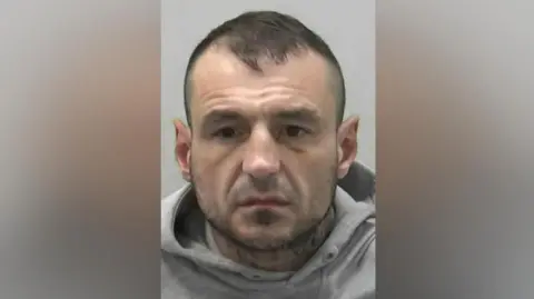 Northumbria Police Mugshot of Tracey. He has short dark hair, a dark stubble beard and is wearing a grey hoodie. 