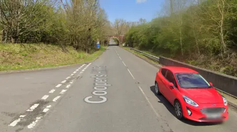 A Google street view of Cooper's Hill Road. There is a red car on the road with greenery on banks either side. There is a tunnel in the distance.