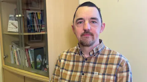 A man with short black hair and goatee beard sitting beside a book case while wearing a checked shirt