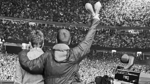 Oasis walk out on stage. The photograph is in black and white and you can see the brothers walking out on stage to a sea of people.