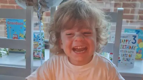 Contributed Wilfred is pictured during his third birthday celebrations. He is smiling with his mouth open and looking away from the camera. He is sat on a chair with a window behind him. Birthday cards have been placed on the windowsill behind him.