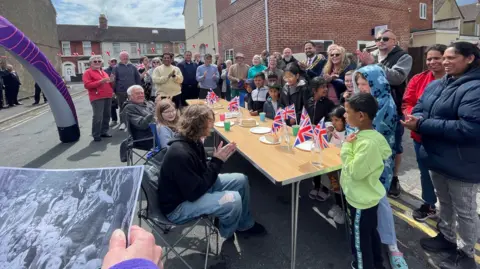 Recreation of a VE Day Street party on a street in Swindon showing children and locals residents and a mayor standing and sitting around trestle tables, surrounded by bunting