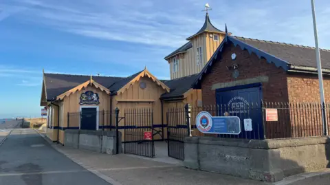 The South Shields Volunteer Life Brigade headquarters, a yellow Victorian building with blue doors. It is at the bottom of the pier which then goes out to sea.