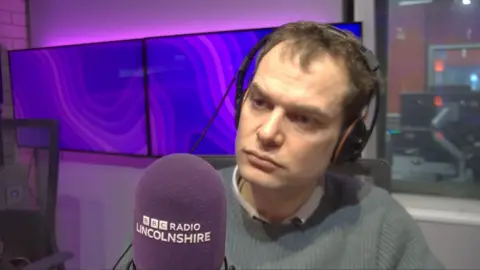 A man with short brown hair who is clean shaven and wearing headphones. He is sat in a purple BBC Radio Lincolnshire studio and is sat next to a purple mic.