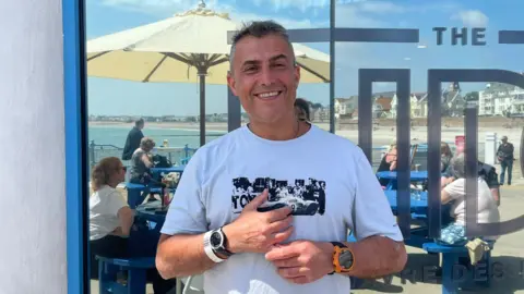 BBC Nelson De Sousa stands in front of a glass window with the partially visible text "THE TID." Behind them is an outdoor seating area with people at blue tables under large umbrellas, and a view of the beach and ocean. He is wearing a white t-shirt with a black graphic design, two watches—one on each wrist—and is holding an object in their hands.