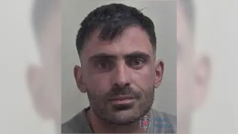 A custody image of Huseyin Kalyoncu. He has short brown hair and stubble. He is looking directly at the camera and is wearing a grey jumper.