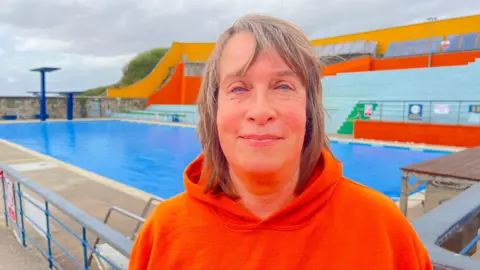 Claudia Amos smiles at the camera. She is wearing an orange hoodie, and in the background is the open-air pool, brightly coloured seating in orange, yellow and blue behind it.