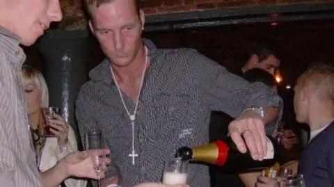 Adam Minett stands in a nightclub in Liverpool, pouring champagne from a bottle in his left hand. He is surrounded by other people. 