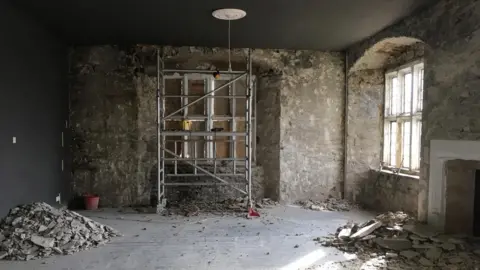 Christine Tallon A stripped back room being renovated. There is piles of plaster on the floor and scaffolding inside the room.