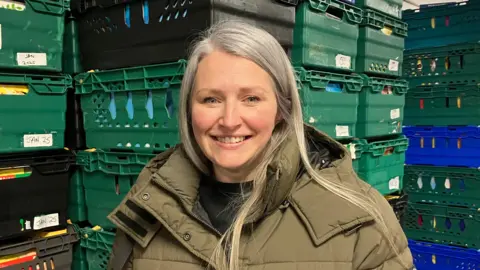 A woman wearing a light brown long jacket stands smiling and looking into the camera. She has long grey hair.  Behind her are boxes of food which are piled high and well above her head.