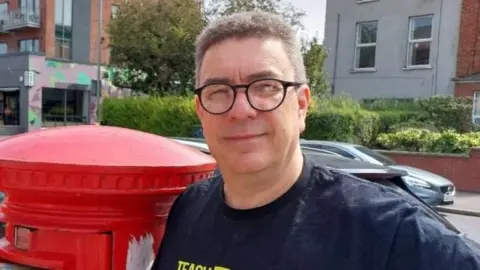A man standing beside a red post box. He has short grey hair and is wearing glasses and a navy t-shirt. 